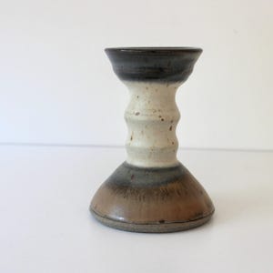 May include: A brown, white and grey ceramic candle holder with a flared base and a textured, cylindrical middle section.
