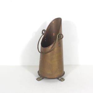 May include: A vintage copper coal scuttle with a curved handle and a flared opening. The scuttle has a conical shape and rests on three small, claw-shaped feet. The metal has a warm, aged patina.