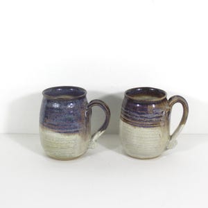 Peut inclure: Deux mugs en céramique faits à la main. Chaque mug a un dessus bleu foncé, un fond beige clair et une anse incurvée. Les mugs sont côte à côte sur un fond blanc. Les mugs sont probablement pour le café ou le thé.