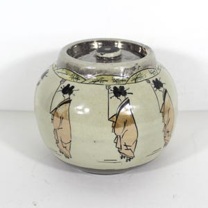 May include: A round ceramic jar with a silver lid, decorated with black and gold painted figures of women in traditional Japanese clothing. The jar has a white background with a subtle green glaze.