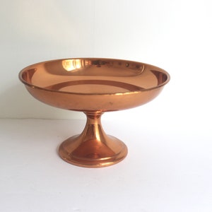 Peut inclure: Un bol sur piédestal de couleur cuivre, avec un large bassin peu profond et une base arrondie et évasée. Le bol a une surface lisse et réfléchissante et est probablement en métal. C'est un objet décoratif.