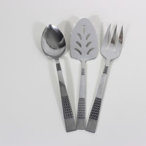 May include: A set of three stainless steel serving utensils: a spoon, a pie server, and a fork. Each utensil has a decorative geometric pattern on the handle. The pie server has a slotted design.