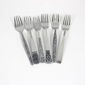 May include: A set of six stainless steel forks arranged side by side. Each fork features a distinct handle design, including floral patterns, geometric shapes, and one plain handle. The tines are silver-toned and polished, showing a variety of styles.