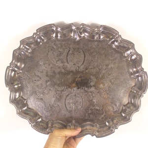 May include: An oval, silver-coloured serving tray with ornate, embossed floral and scrollwork designs. The tray has a scalloped edge and a slightly tarnished appearance. A hand is visible holding the tray.