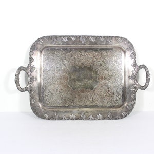 May include: A silver serving tray with ornate detailing and two handles. The tray is rectangular in shape and has a slightly raised rim.