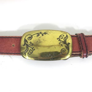 May include: A brown leather belt with a gold-toned buckle featuring a decorative floral design.