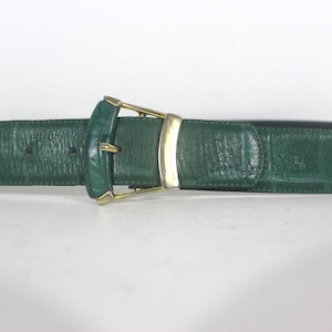 May include: A green leather belt with a gold-coloured buckle. The belt has a textured surface and a dark green colour. The buckle is rectangular with a gold-coloured frame.