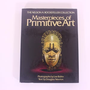May include: A hardback book titled "Masterpieces of Primitive Art" from The Nelson A. Rockefeller Collection. The cover displays a golden mask against a dark background. The book's text is by Douglas Newton and photographs by Lee Boltin.