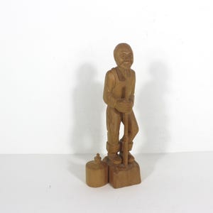 May include: A carved wooden figurine of a man in overalls holding a tool, standing on a wooden base. A small, cylindrical wooden object sits to the left of the figure. The wood has a light brown colour.