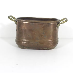 May include: An oval copper planter with two brass handles. The planter has a worn, aged look and is perfect for adding a rustic touch to your home decor.
