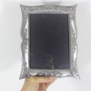 May include: A silver picture frame with a floral design and two intertwined hearts at the top. The frame is designed to hold a 10x15 cm photo.
