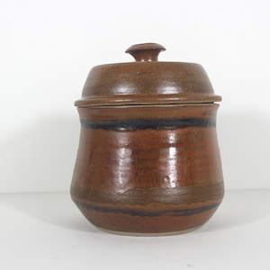 May include: A brown ceramic jar with a lid. The jar has a wide mouth and a rounded body. It has a brown glaze with black stripes around the middle.