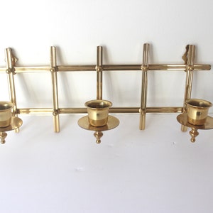 May include: A gold metal wall-mounted candle holder with three candle cups. The holder has a geometric design with a rectangular frame and vertical bars.