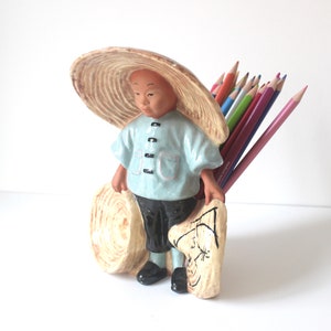 Peut inclure: Un porte-crayons en céramique en forme de personnage portant un chapeau de paille et des vêtements traditionnels. La figurine tient une charrette en bois remplie de crayons de couleur. Le support est peint en bleu clair, noir et beige.