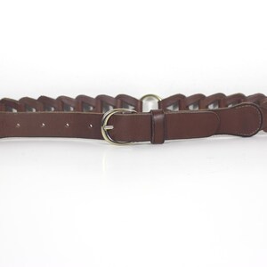 Peut inclure: Une ceinture en cuir marron avec un motif tressé et une boucle dorée. La ceinture a un style classique et convient à diverses tenues. La ceinture est présentée sur un fond blanc.