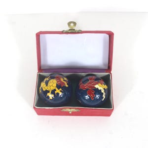 May include: A pair of dark blue spherical meditation balls featuring colorful dragon and phoenix designs in yellow, red, and white. The two balls are housed in a rectangular red velvet-lined storage box with a gold-toned metal latch closure.