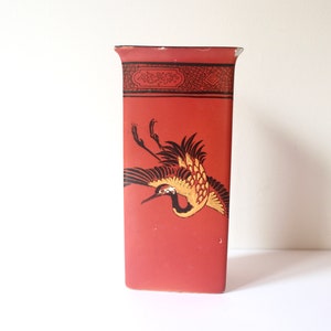 May include: A red ceramic vase with a black and gold design of a crane in flight. The vase has a rectangular shape and a simple, elegant design.