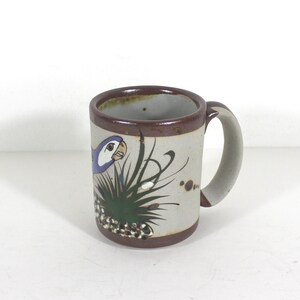 May include: A brown ceramic mug with a white glaze and a painted design of a blue and purple parrot in a green bush. The mug has a brown handle.