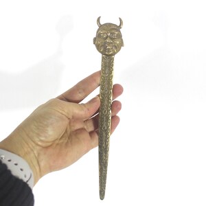 May include: A gold-coloured metal letter opener with an intricate design. The handle is shaped like a devil's head with horns.