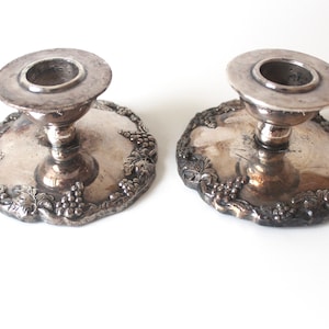 May include: A pair of antique silver candle holders. Each features a round base with a grape vine pattern. They have a short stem and a circular top for a candle. The silver has a tarnished look, typical of aged items.