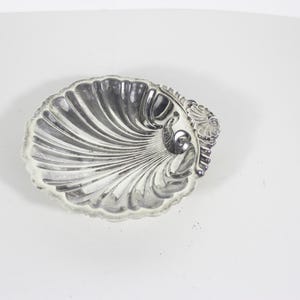 Peut inclure: Un plat en métal argenté en forme de coquille Saint-Jacques. Le plat présente un centre concave profond avec des crêtes cannelées rayonnantes qui s'étendent d'un point central vers le bord festonné. Un petit détail décoratif est visible.