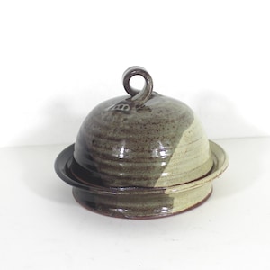 May include: A handmade ceramic butter dish with a lid. The dish is glazed in shades of green, brown, and white. The lid has a unique, twisted handle.