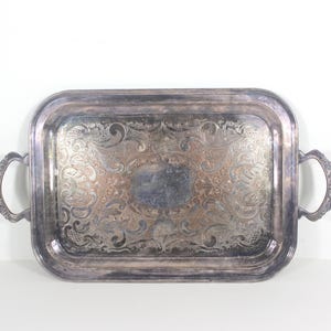 May include: A silver serving tray with ornate embossed designs and two handles. The tray is rectangular and has a slightly raised edge.