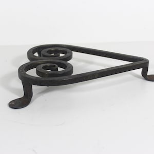 May include: A black metal heart-shaped trivet with two decorative swirls. The trivet has three legs.