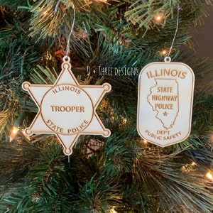 May include: Two wooden Christmas ornaments featuring the Illinois State Police logo. One ornament is a star-shaped badge with the text "Illinois State Police Trooper" and the other is a rectangular ornament with the text "Illinois State Highway Police Dept. Public Safety".