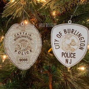 May include: Two wooden Christmas ornaments hanging on a tree. One is a police badge with "Captain Burlington Police" and "9066" engraved. The other is a shield-shaped ornament with "City of Burlington Police NJ" engraved.