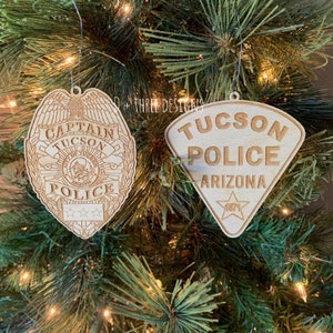 May include: Two wooden Christmas ornaments hanging on a green Christmas tree. One ornament is a police badge with the words "CAPTAIN TUCSON POLICE" and stars. The other is a badge-shaped ornament with "TUCSON POLICE ARIZONA" and a star.