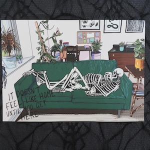 May include: An illustration depicting two skeletons embracing on a green sofa in a domestic setting. The skeletons are in a loving embrace. The text "IT DOESN'T FEEL LIKE HOME UNTIL YOU GET HERE" is printed on the bottom left corner of the image.