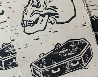May include: A stack of black and white prints featuring a skull and a coffin. The prints are on a textured paper.