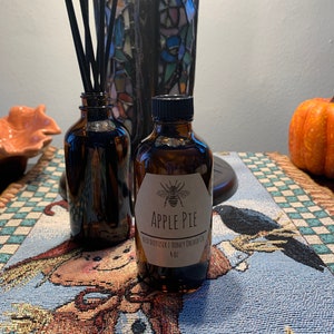 May include: A brown glass bottle with a black lid and a white label that says "Apple Pie Reed Diffuser | Honey Orchid Co. 4 oz". The label also features a black and white illustration of a bee. The bottle is sitting on a blue and white checkered tablecloth.