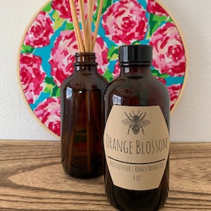 May include: A brown glass bottle with a brown paper label that reads "Orange Blossom Reed Diffuser | Honey Orchid Co. 4 oz". The label features a black and white illustration of a bee. The bottle is sitting on a wooden surface with a round, pink and blue floral embroidery hoop behind it.