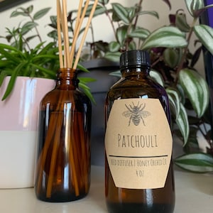 May include: A brown glass bottle with a brown label that says "Patchouli Reed Diffuser | Honey Orchid Co. 4 oz". The label also features a black and white illustration of a bee. The bottle is filled with a clear liquid and has wooden sticks in it.