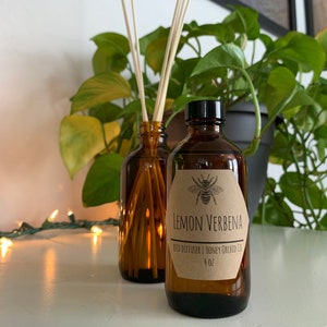 May include: Two amber glass reed diffusers with wooden reeds. One bottle has a label that reads "Lemon Verbena" with a bee illustration. The bottles are 4 oz. each. A green plant is in the background.