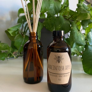 May include: Two brown glass bottles with a reed diffuser and a brown paper label. The label has a bee illustration and the text "Macintosh Apple Reed Diffuser | Honey Orchid Co. 4 oz."