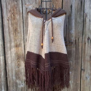May include: A hooded poncho featuring a two-tone knit design with a beige upper section and a dark brown lower section. The garment includes a hood with brown drawstring cords ending in wooden beads, and a long fringe trim along the bottom edge.