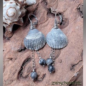 Könnte beinhalten: Handgefertigte Ohrringe mit grauen Muscheln und silbernen Drahtdetails. Jeder Ohrring hat eine Kette mit drei grauen Perlen. Die Ohrringe werden auf einer Holzoberfläche präsentiert. Der Text "zwischenwelt" ist sichtbar.