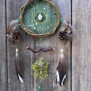 May include: Green, brown and white dreamcatcher with feathers, pine cones, beads and a bell hanging from a wooden base. The dreamcatcher is decorated with a spider and moss.