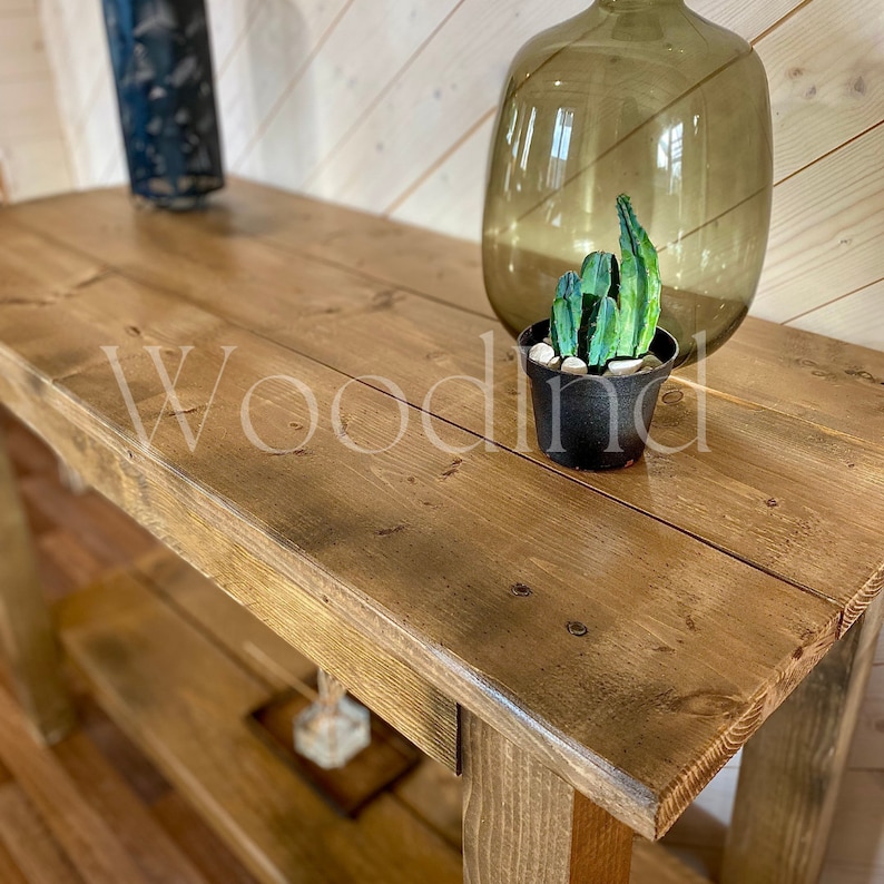May include: A rustic, wooden console table with a lower shelf. The tabletop is made of wide, stained planks. A decorative glass bottle, a small potted cactus, and a dark metal lantern are on the table. The word "Woodind" is printed on the table.