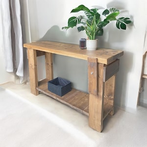 Peut inclure: Une console en bois rustique avec une étagère inférieure. La table a une finition en bois naturel et est faite de bois de récupération. La table mesure 122 cm de long et 41 cm de profondeur.
