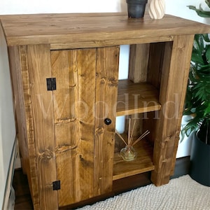 Könnte beinhalten: Ein rustikaler Holzschrank mit einer einzigen Tür und zwei Regalen. Der Schrank ist in einem dunklen Braunton gebeizt und hat schwarze Metallbeschläge und einen Knauf. Der Schrank steht auf einem weißen Teppich.