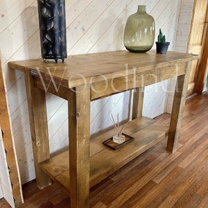 May include: A rustic, wooden console table with a lower shelf. The table is made of light brown wood and has a rectangular top. A black metal lamp, a green glass vase, and a small potted cactus are on the tabletop. The table is in a room with wood paneling.