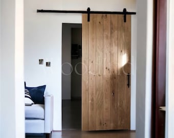 May include: A natural wood barn door with vertical planks, mounted on a black metal track. The door is open, revealing a glimpse of a living space with a white sofa and blue pillows. The door's hardware includes a black handle.