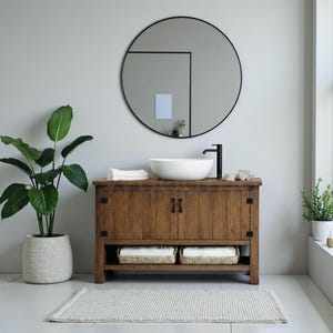 Puede incluir: Un mueble de baño rústico de madera con un lavabo blanco y un grifo negro. Un gran espejo redondo cuelga sobre el mueble. Dos cestas con toallas dobladas están en el estante inferior. Una planta en maceta y una alfombra completan la decoración.