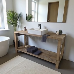 May include: A rectangular wooden bathroom vanity featuring a light brown finish, a lower storage shelf, and a white ceramic vessel sink with a matte black faucet. The vanity is topped with a white sink and holds dark gray folded towels.