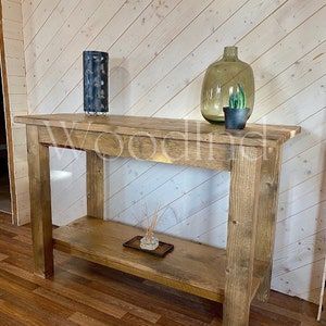 May include: A rustic, wooden console table with a lower shelf. The table is stained a warm brown and features a rectangular top. Decorative items include a blue metal lantern, a large glass vase, and a small potted cactus. The word "Woodland" is partially visible.