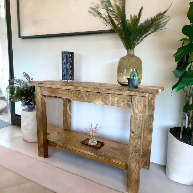 May include: A rustic, wooden console table with a lower shelf. The table is made of light brown wood and has a natural, weathered appearance. A decorative vase, a glass bottle, and a small potted cactus are displayed on the table.